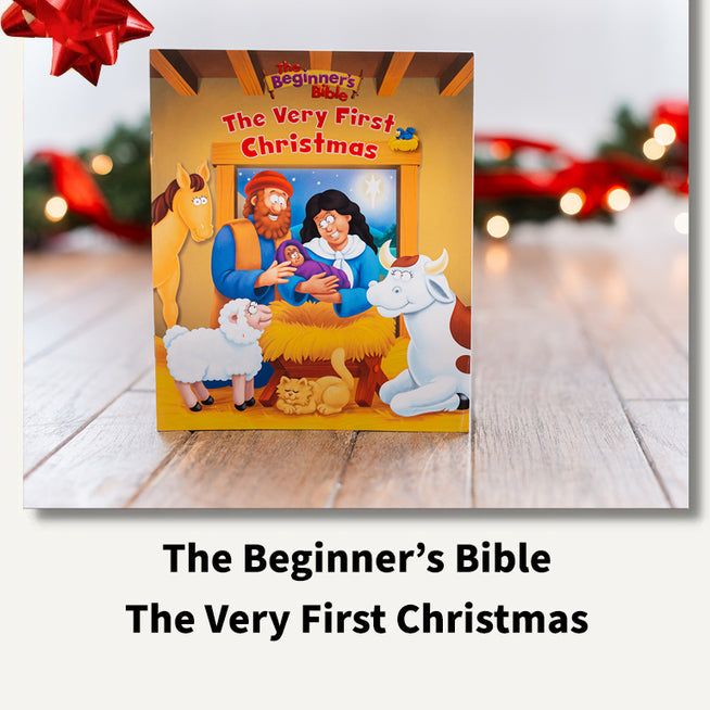 The Beginner's Bible: The Very First Christmas book cover displayed on a light wood surface with a blurred festive background and a red gift bow graphic in the top left. Text below reads 'The Beginner's Bible The Very First Christmas'.